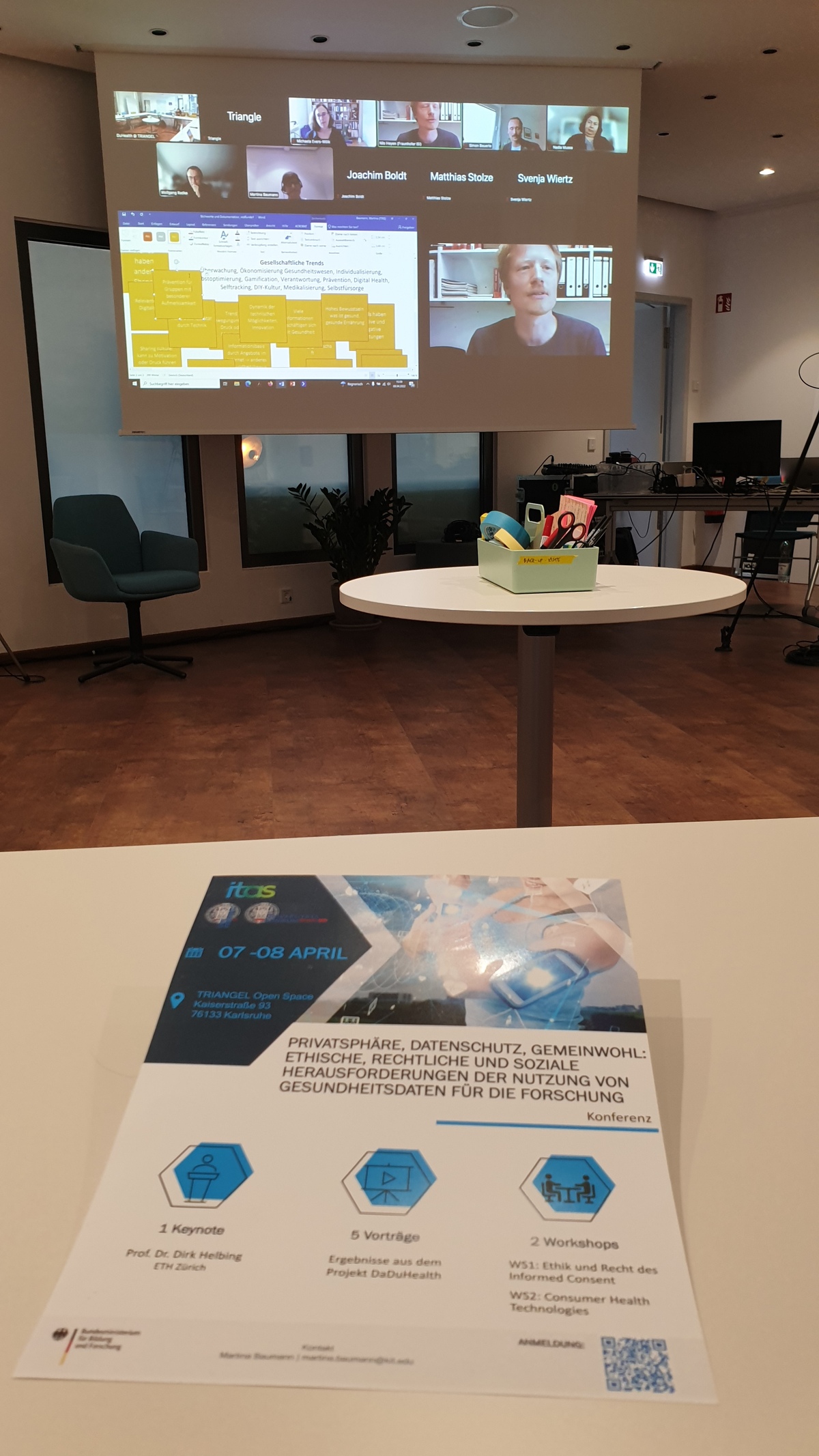Im Vordergrund liegt das Workshop-Programmheft auf einem Tisch, im Hintergrund sieht man ein aktives Online-Meeting, das auf eine Leinwand projiziert wird.