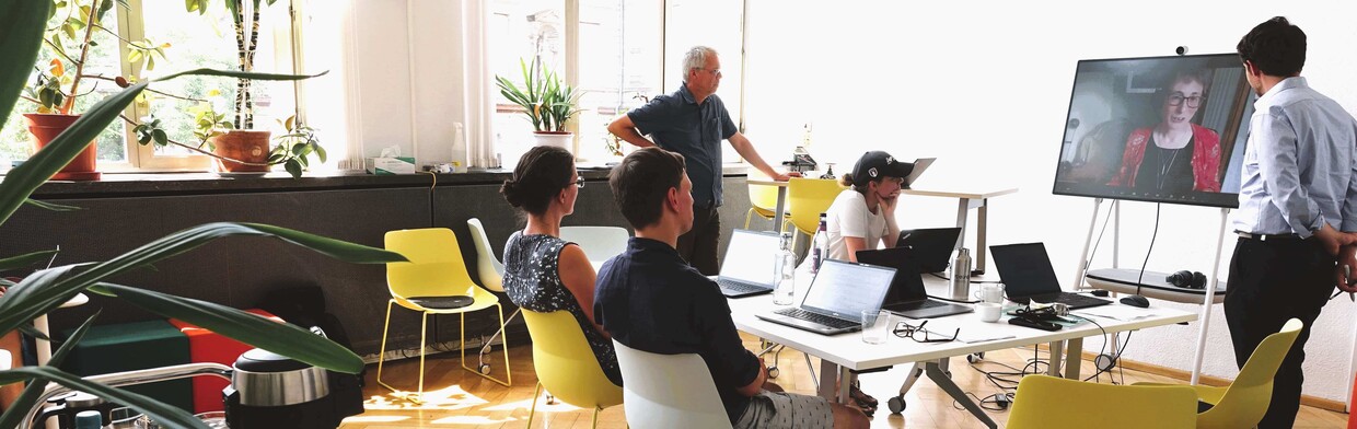 People sitting and standing around a table, talking to a person on a screen.